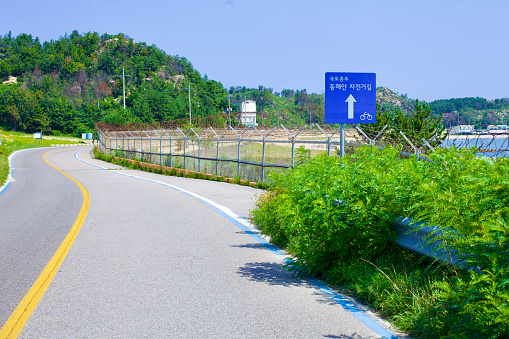 Goseong County, South Korea - July 30, 2019: Alongside a road near the north end of Gonghyeonjin Beach, a bike path sign and a distinct blue line indicate the route as part of the Gangwon bike path, with barbed wire-topped fencing separating the path from the coastal area.