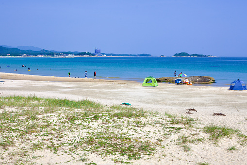Goseong County, South Korea - July 30, 2019: On a luminous summer day, Sampo Beach unfolds its flat sandy shores into the blue waters, dotted with tents and people enjoying floats or wading, while flat rocks and seaside islands create a serene background.