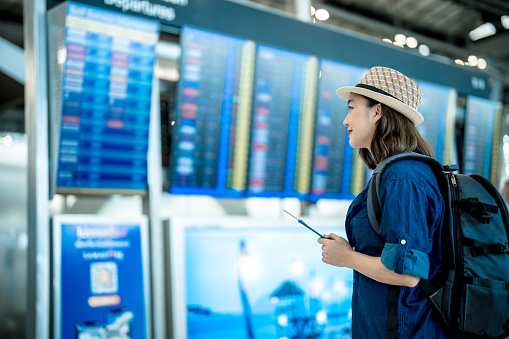 Young asian woman with a backpack holding passport and boarding pass as a hand in international airport looking at the flight information board, checking her flight