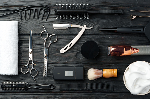 A variety of professional barber tools are neatly arranged on a dark wooden surface, including scissors, a straight razor, combs, clippers, and grooming products, suggesting preparation for a grooming session.