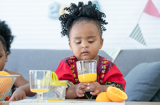 African cute kid girl drinking fresh oranges juice from glass at home for breakfast after making freshly squeezed orange juice on manual juicer. Healthy lifestyle and learning concept