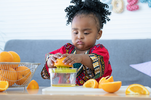 African cute kid girl squeezing fresh oranges at home. Adorable child with funny face expression makes preparing freshly squeezed orange juice on manual juicer. Healthy lifestyle and learning concept