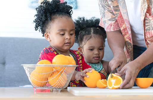 Little African cute daughters helping her senior father to slice orange on cutting board and preparing food at home. Children siblings enjoy pick fruit from basket to give father cut for breakfast