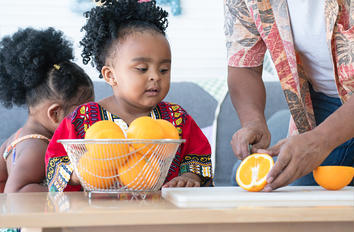 Little African cute kid girl helping her senior father to cut orange on cutting board and preparing food at home