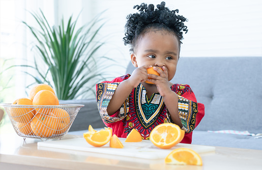 African cute kid girl have fun and enjoy eating fresh orange for breakfast at home. Adorable child eating sliced orange in hands with messy mouth. Healthy lifestyle and learning concept