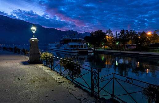 Annecy. France. September 24, 2019. View of Lake Annecy from the embankment at dusk in the early morning. There is a street lamp, a boat, mountains and a bright pre-dawn sky.