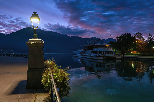 Annecy. France. September 24, 2019. Street lamp on the embankment at dusk in the early morning. There is a ship, mountains and a bright pre-dawn sky with dark clouds.