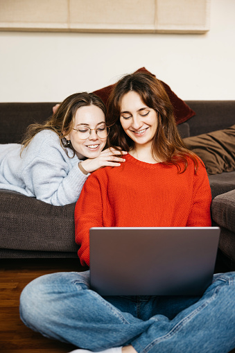 Lesbian smiling couple sitting together on a sofa at home with a laptop. Young female gay couple having fun using a laptop in an intimate and cozy atmosphere.