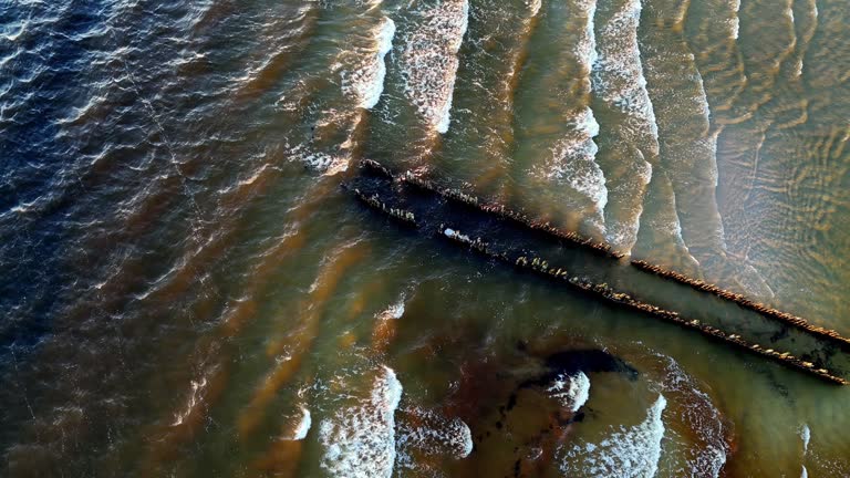 aerial view of a long pier jutting out into a turquoise ocean with white-capped waves crashing against the shore, with a few old boats bobbing nearby