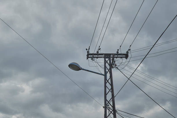Street light mounted on a metal electrical post, accentuated by intertwining power lines against overcast sky. Selective focus.