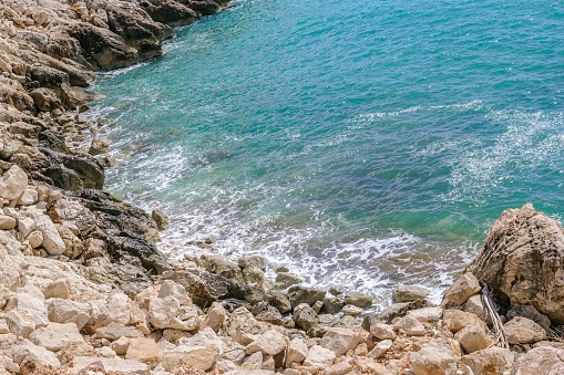 A picturesque rocky beach in the Mediterranean Sea with clear blue water and stones. The beach is in a cove surrounded by cliffs. Crystal clear water allows you to see the stones on the seabed.