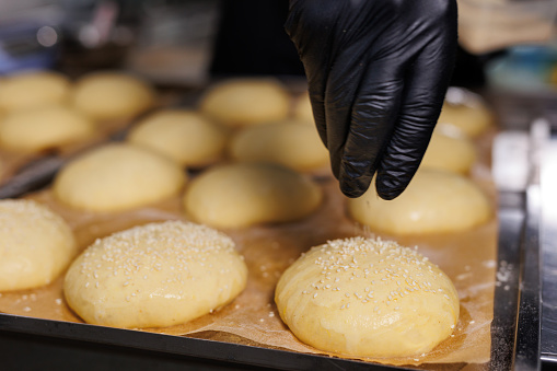 Close-up of the chef's gloved hands, who sprinkles sesame seeds on burger buns before baking