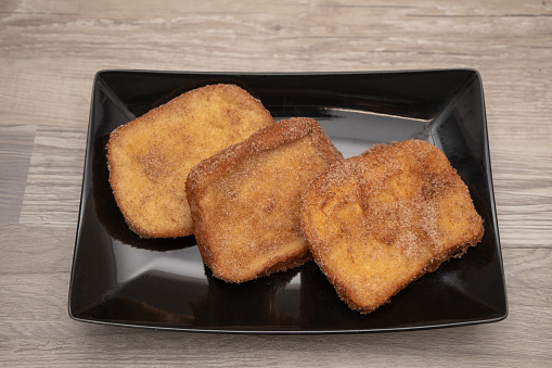Three golden torrijas dusted with sugar and cinnamon on a sleek black serving plate, ready to be enjoyed.