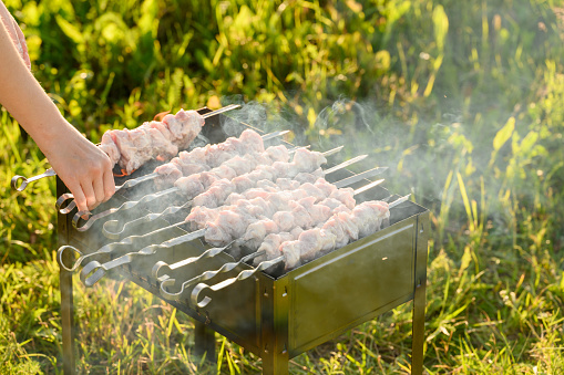 Woman's hand places pieces of fresh raw shish kebab, strung on skewers, on the grill with coals and smoke. Cooking food on coals. Family picnic, summer vacation.