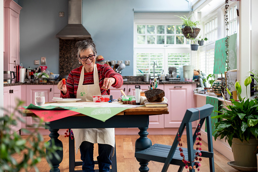 A three-quarter-length shot of a mature adult female in the kitchen of her home in Hexham, Northumberland. She is sitting at a table which is laid with art and crafts equipment, enjoying a print-making session using wooden blocks, paint and paper. She is wearing casual clothing and an apron.

Videos of this scenario available.