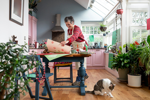 A wide full-length shot of a mature adult female in the kitchen of her home in Hexham, Northumberland. She is standing at a table which is laid with art and crafts equipment, enjoying a print-making session using wooden blocks, paint and paper. She is smiling, wearing casual clothing and an apron. Her pet cat is sitting on the floor beside the table, licking its leg.
