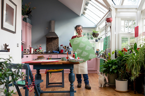 A wide full-length shot of a mature adult female in the kitchen of her home in Hexham, Northumberland. She is standing beside a table which is laid with art and crafts equipment, proudly holding up a piece of paper with a patterned printed design on that she has created. She is smiling, wearing casual clothing and an apron. Her pet cat is sitting on the floor beside her.