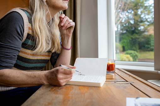 A table-height side-view shot of an unrecognisable adult female sitting at a desk in a window in a home in Hexham, Northumberland. She is holding a pen and there is a journal and burning candle on the table in front of her. She is wearing casual clothing and outside there is greenery visible.