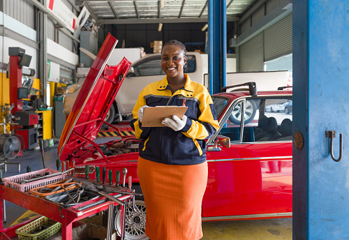 Young woman with a work uniform and protective glove standing in an automotive workshop or garage, smile while holding a clipboard. A red car with an open hood repairing in the background.