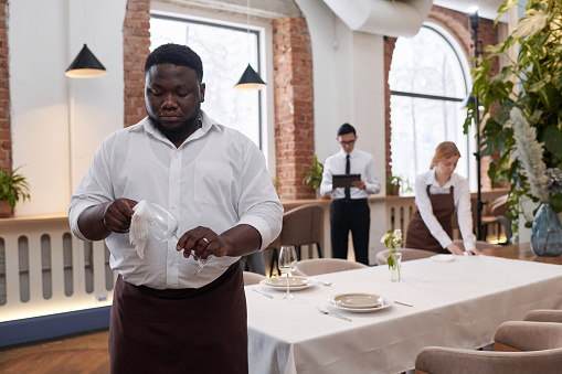 Selective focus shot of young Black waiter working in modern restaurant polishing wine glass, copy space