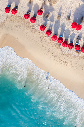 Rows of red umbrellas line the sandy beach, casting distinct, long shadows. Two people wade in the clear blue water near the shore as gentle waves crash onto the sand. The image is taken from an aerial perspective.