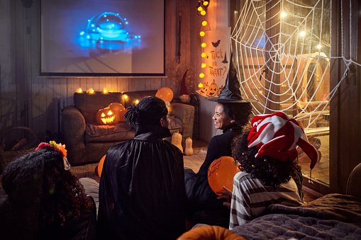 Back view of happy African American parents talking while watching a move with their kids on TV during Halloween night at home.