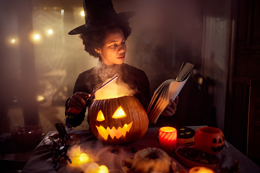 African American woman in witch costume reading a book while casting spells on smoking Jack O' Lantern during Halloween at home.