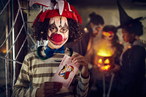Portrait of happy African American boy in clown costume holding Joker card on Halloween at home and looking at camera. There are people in the background.