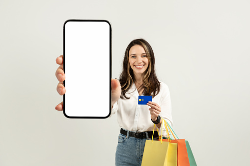 Happy european young woman showing a smartphone with a blank screen and holding a credit card with colorful shopping bags, suggesting online shopping on a light background