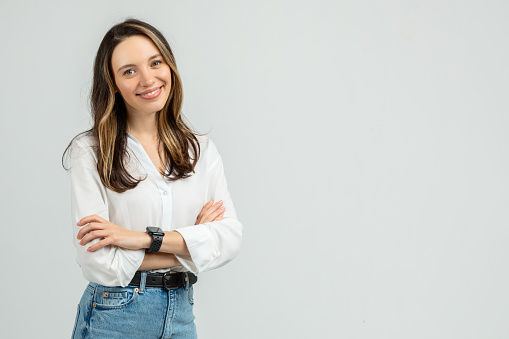 A confident young european woman with long brunette hair smiles gently at the camera, arms crossed, dressed in a white blouse and blue jeans, with a smartwatch on her wrist