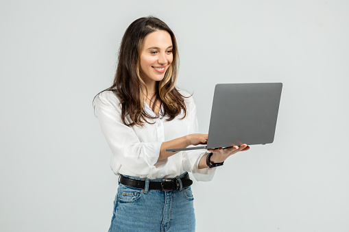 Engaged young woman in a white blouse and blue jeans focused on a grey laptop she is holding, with a subtle smile and a smartwatch on her wrist, standing against a grey backdrop