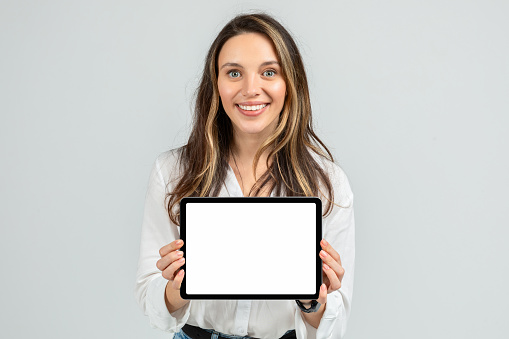 Enthusiastic young woman holding up a tablet with a blank screen, wearing a white blouse and jeans, with a beaming smile and a smartwatch, against a plain grey background