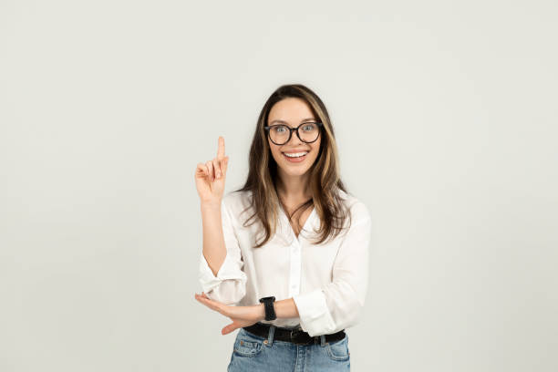 istock Enthusiastic young woman with glasses cheerfully pointing upwards 2103843019