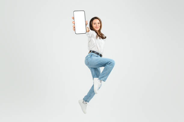 istock Dynamic young woman in mid-air jump, joyfully presenting a smartphone with a blank screen 2103843021