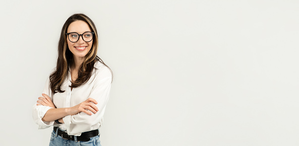 Smiling european woman with glasses standing with arms crossed, dressed in a white blouse and blue jeans, exuding confidence and professionalism on a clean background, studio