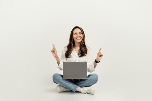 Happy european woman sitting cross-legged on the floor with a laptop, pointing upwards, wearing a white blouse and jeans, looking inspired with a bright idea, studio. Business, work