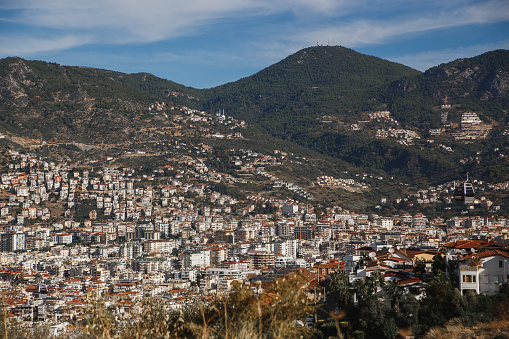 A city nestled in a natural landscape surrounded by a majestic mountain range, with a single towering mountain in the background under a sky filled with fluffy clouds