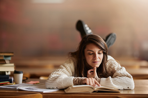 Smiling female student reading a book while relaxing on a desk in library.