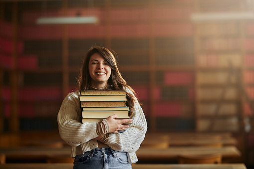 Portrait of happy college student standing in library with stack of books and looking at camera.