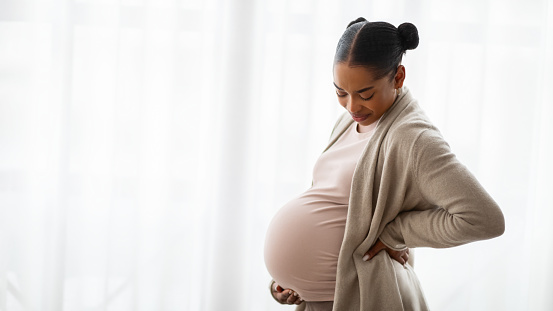 Maternity, pregnancy concept. Beautiful young african american pregnant woman wearing casual clothing standing next to window at home, embracing her big tummy, panorama with copy space