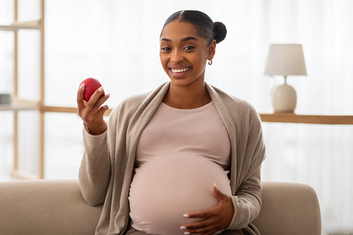 Positive pretty young african american expecting woman sitting on couch at home, embracing her big tummy, holding red apple and smiling at camera. Healthy nutrition during pregnancy