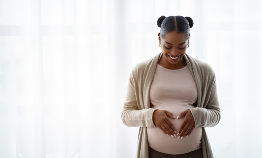 Smiling young black pregnant woman wearing comfy clothing holding hands on her big belly, enjoying her pregnancy, expecting baby, standing by window at home, panorama with copy space