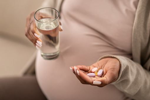 Cropped of black pregnant woman holding medicine drugs and glass of water, taking vitamins or medication for expectants, sitting on couch at home. Health care during pregnancy concept