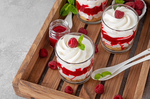 Raspberry trifle with whipped cream, berry sauce, cookie crumbs and mint leaves in a glass on a wooden tray. Summer diet dessert