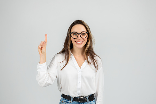 Confident glad european young woman with glasses pointing upwards, wearing a white blouse and blue jeans, standing against a grey background with a positive expression, studio