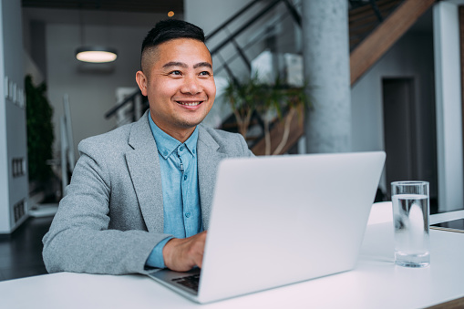 Shot of a businessman sitting on desk in modern office and working on laptop. Portrait of an elegant handsome asian businessman sitting in his office and using laptop.