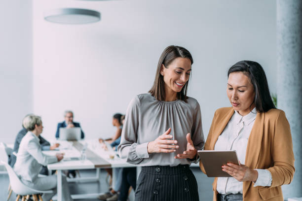 Business people in the office. Shot of two female coworkers having a discussion in modern office. Businesswomen in meeting using digital tablet and discussing business strategy. Confident business people working together in the office. Corporate business persons discussing new project and sharing ideas in the workplace. Successful businesswomen standing with their colleagues working in background. business stock pictures, royalty-free photos & images
