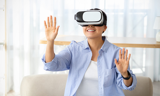 Joyful asian young woman wearing modern VR glasses while sitting on couch in living room, touching blank space, chinese lady experiencing virtual reality session at home