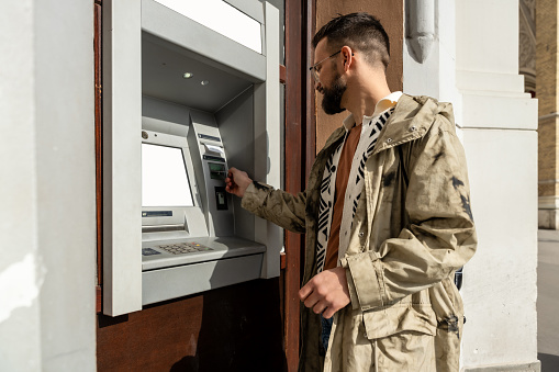 Young fashionable hipster backpacker traveler man using credit card to withdraw money from ATM machine on the street in the city for life in new country and city. Modern lifestyle of male person