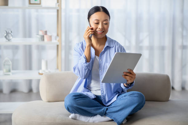 Chinese businesswoman working from home, have phone conversation Chinese millennial businesswoman working from home, sitting on couch, using digital tablet, have phone conversation with business partner or client, smiling, copy space. Entrepreneurship project manager remote stock pictures, royalty-free photos & images